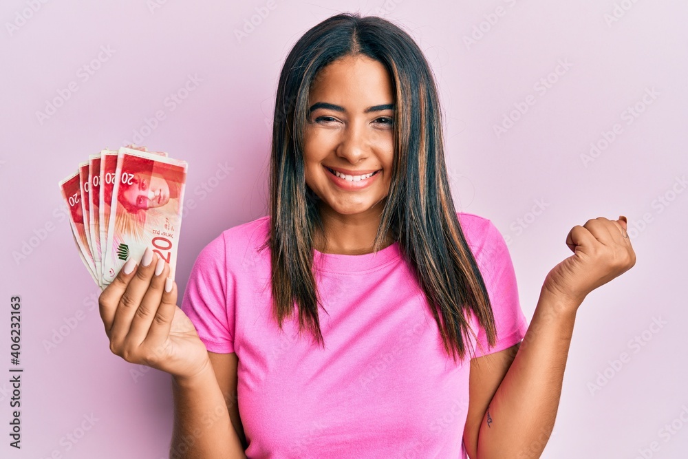 Young latin girl holding 20 israel shekels banknotes screaming proud, celebrating victory and success very excited with raised arm