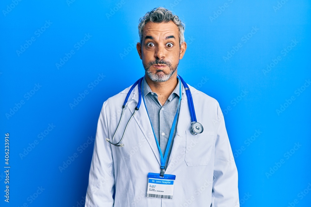 Middle age grey-haired man wearing doctor uniform and stethoscope puffing cheeks with funny face. mouth inflated with air, crazy expression. Middle age grey-haired man wearing doctor uniform and stethoscope puffing cheeks with funny face. mouth inflated with air, crazy expression.