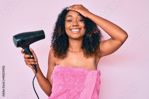 Young african american woman wearing shower towel holding dryer stressed and frustrated with hand on head, surprised and angry face