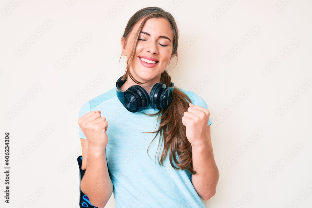 Beautiful young caucasian woman wearing gym clothes and using headphones very happy and excited doing winner gesture with arms raised, smiling and screaming for success. celebration concept. Beautiful young caucasian woman wearing gym clothes and using headphones very happy and excited doing winner gesture with arms raised, smiling and screaming for success. celebration concept.