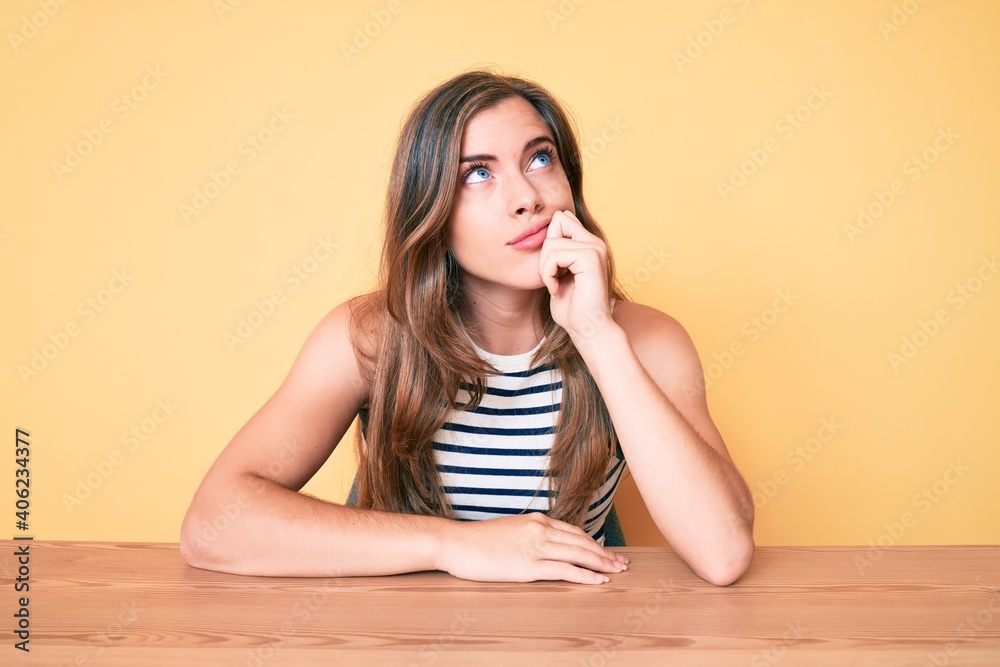 Beautiful young caucasian woman wearing casual clothes sitting on the table serious face thinking about question with hand on chin, thoughtful about confusing idea Beautiful young caucasian woman wearing casual clothes sitting on the table serious face thinking about question with hand on chin, thoughtful about confusing idea