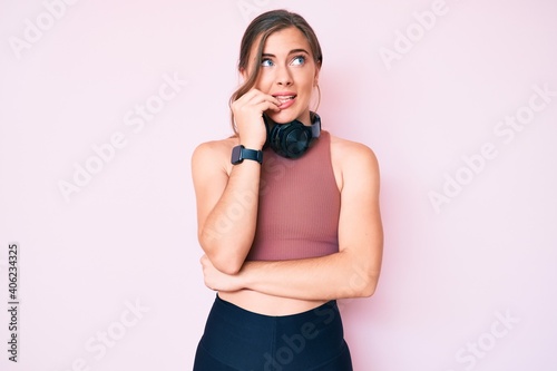 Beautiful young caucasian woman wearing gym clothes and using headphones looking stressed and nervous with hands on mouth biting nails. anxiety problem.