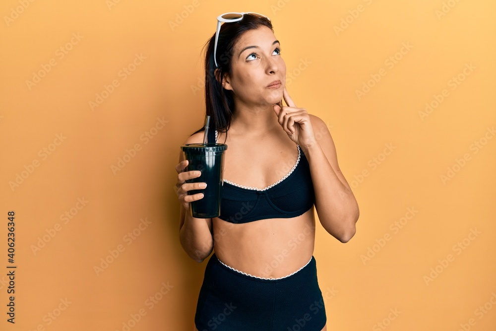 Young latin woman wearing bikini drinking soda serious face thinking about question with hand on chin, thoughtful about confusing idea
