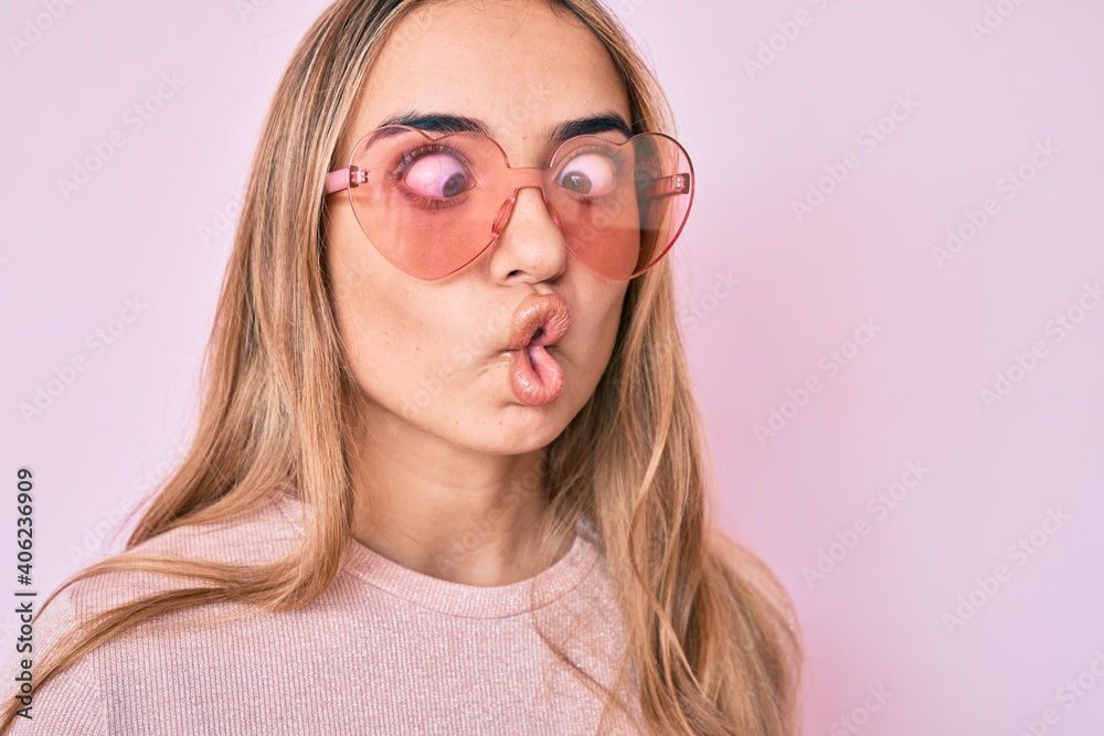 Young beautiful blonde woman wearing heart shaped sunglasses making fish face with mouth and squinting eyes, crazy and comical.