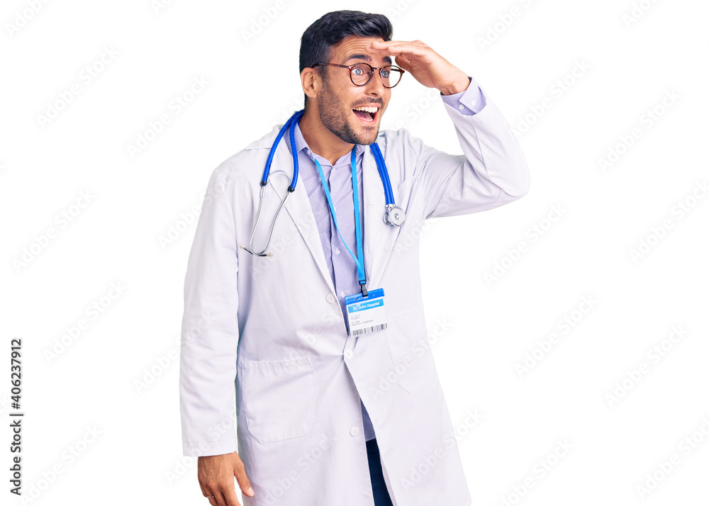 Young hispanic man wearing doctor uniform and stethoscope very happy and smiling looking far away with hand over head. searching concept.
