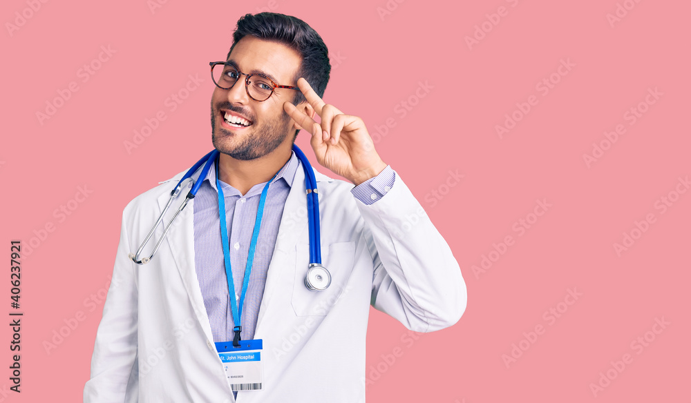 Young hispanic man wearing doctor uniform and stethoscope doing peace symbol with fingers over face, smiling cheerful showing victory Young hispanic man wearing doctor uniform and stethoscope doing peace symbol with fingers over face, smiling cheerful showing victory
