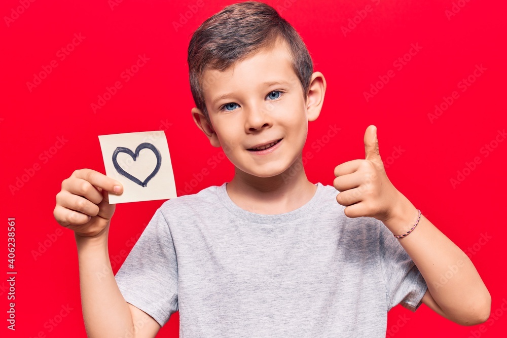 Cute blond kid holding heart reminder smiling happy and positive, thumb up doing excellent and approval sign Cute blond kid holding heart reminder smiling happy and positive, thumb up doing excellent and approval sign