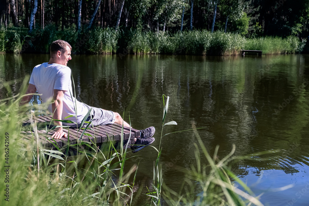 Happy relaxed man sitting on wooden pier on a lake enjoying a sunny summer day surrounded by greenery and trees. Mental health, reboot Happy relaxed man sitting on wooden pier on a lake enjoying a sunny summer day surrounded by greenery and trees. Mental health, reboot