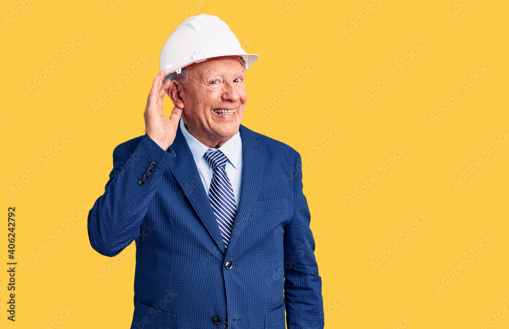 Senior handsome grey-haired man wearing suit and architect hardhat smiling with hand over ear listening an hearing to rumor or gossip. deafness concept.
