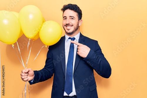 Young hispanic man wearing suit holding balloons smiling happy pointing with hand and finger