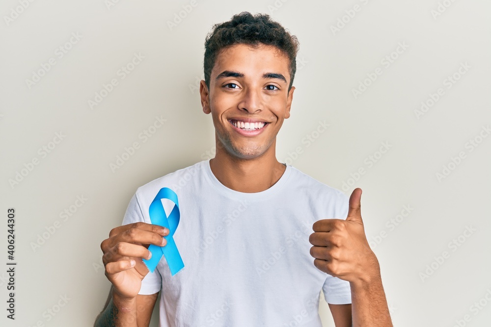 Young handsome african american man holding blue ribbon smiling happy and positive, thumb up doing excellent and approval sign