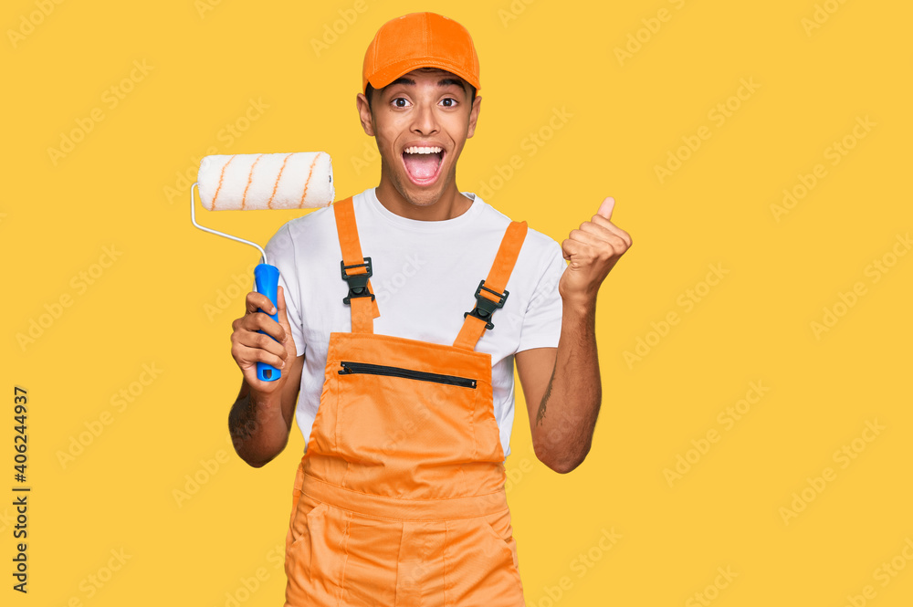 Young handsome african american man wearing cap and painter clothes holding painting roll screaming proud, celebrating victory and success very excited with raised arms Young handsome african american man wearing cap and painter clothes holding painting roll screaming proud, celebrating victory and success very excited with raised arms