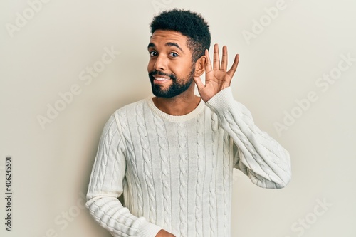 Handsome hispanic man with beard wearing casual winter sweater smiling with hand over ear listening and hearing to rumor or gossip. deafness concept.