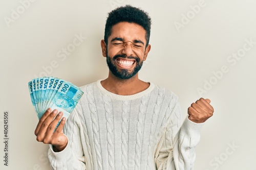 Handsome hispanic man with beard holding 100 brazilian real banknotes screaming proud, celebrating victory and success very excited with raised arm