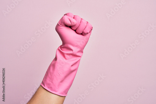 Hand of caucasian young man with cleaning glove over isolated pink background doing protest and revolution gesture, fist expressing force and power