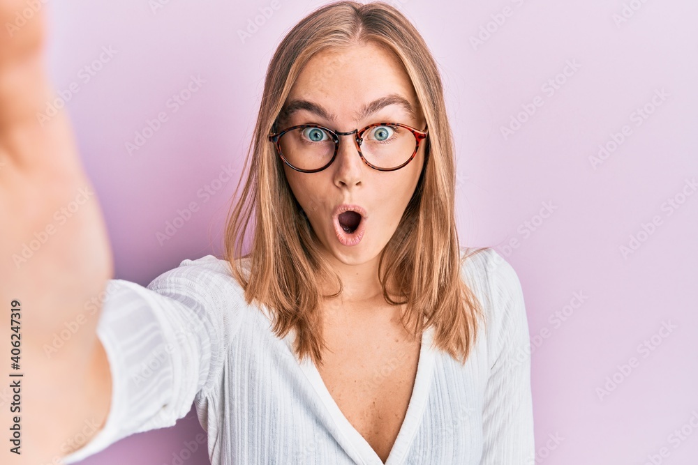 Beautiful blonde woman taking a selfie photo wearing glasses scared and amazed with open mouth for surprise, disbelief face