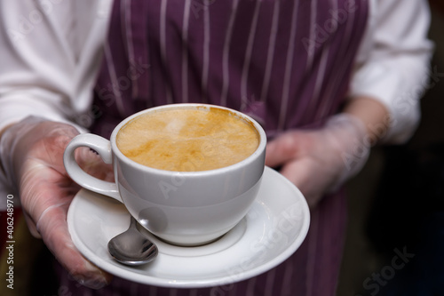 The waiter's gloved hands hold a cup of freshly brewed coffee with foam. Cup close-up. Selective focus.