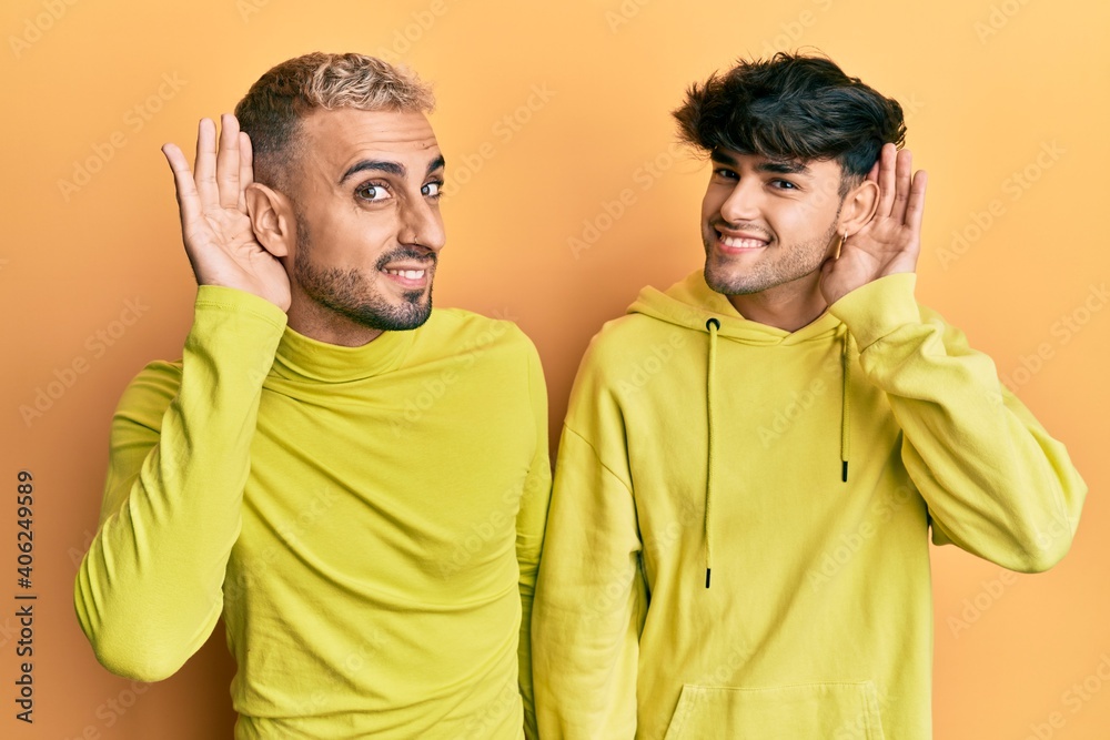 Homosexual gay couple standing together wearing yellow clothes smiling with hand over ear listening an hearing to rumor or gossip. deafness concept.