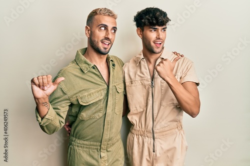 Homosexual gay couple standing together wearing casual jumpsuit smiling with happy face looking and pointing to the side with thumb up.