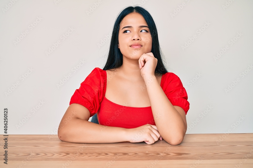 Young beautiful asian girl wearing casual clothes sitting on the table serious face thinking about question with hand on chin, thoughtful about confusing idea Young beautiful asian girl wearing casual clothes sitting on the table serious face thinking about question with hand on chin, thoughtful about confusing idea