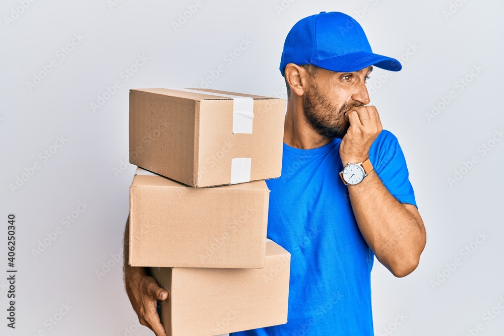 Handsome man with beard wearing courier uniform holding delivery packages looking stressed and nervous with hands on mouth biting nails. anxiety problem.