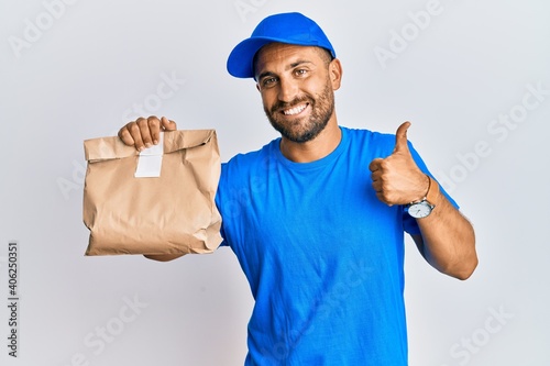 Handsome man with beard holding delivery package smiling happy and positive, thumb up doing excellent and approval sign
