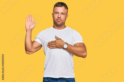 Handsome muscle man wearing casual white tshirt swearing with hand on chest and open palm, making a loyalty promise oath