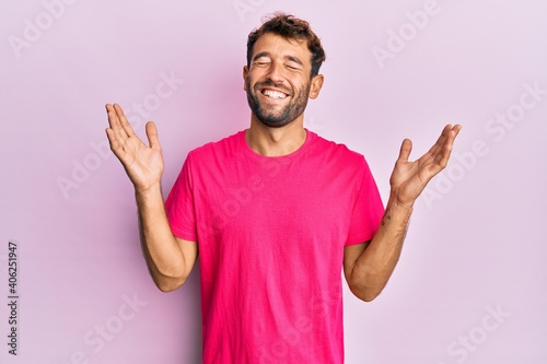 Handsome man with beard wearing casual pink tshirt over pink background celebrating mad and crazy for success with arms raised and closed eyes screaming excited. winner concept