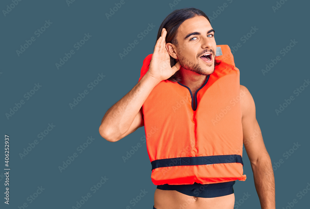 Young handsome man wearing nautical lifejacket smiling with hand over ear listening an hearing to rumor or gossip. deafness concept.