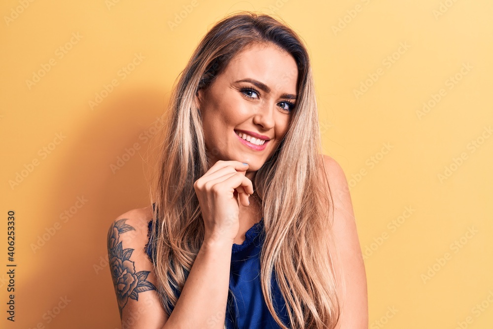 Young beautiful blonde woman wearing casual t-shirt standing over isolated yellow background smiling looking confident at the camera with crossed arms and hand on chin. Thinking positive. Young beautiful blonde woman wearing casual t-shirt standing over isolated yellow background smiling looking confident at the camera with crossed arms and hand on chin. Thinking positive.
