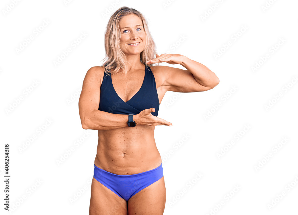 Middle age fit blonde woman wearing bikini gesturing with hands showing big and large size sign, measure symbol. smiling looking at the camera. measuring concept.