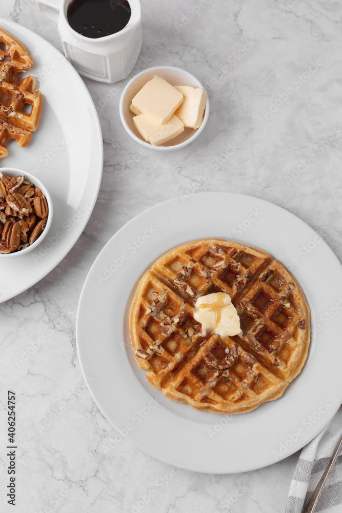 Homemade Pecan Waffles on a White Plate with a Small Dish of Pecans; One Pecan Waffle on a White Plate with Butter and Maple Syrup; Butter and Maple Syrup in Background Homemade Pecan Waffles on a White Plate with a Small Dish of Pecans; One Pecan Waffle on a White Plate with Butter and Maple Syrup; Butter and Maple Syrup in Background
