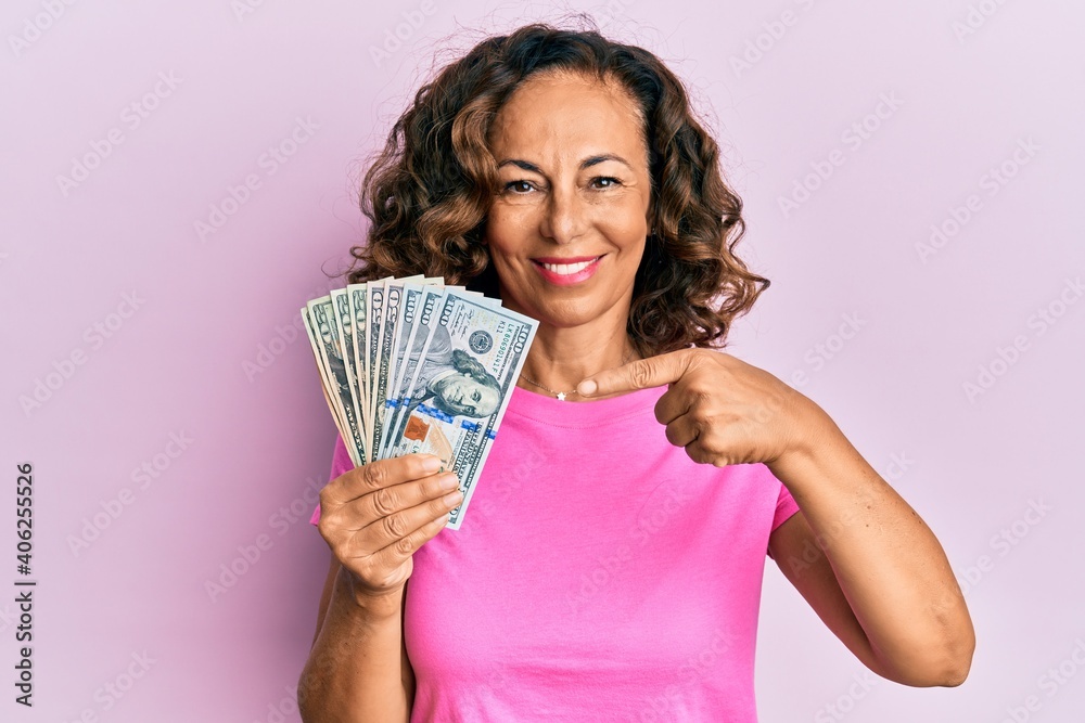 Middle age hispanic woman holding dollars smiling happy pointing with hand and finger Middle age hispanic woman holding dollars smiling happy pointing with hand and finger