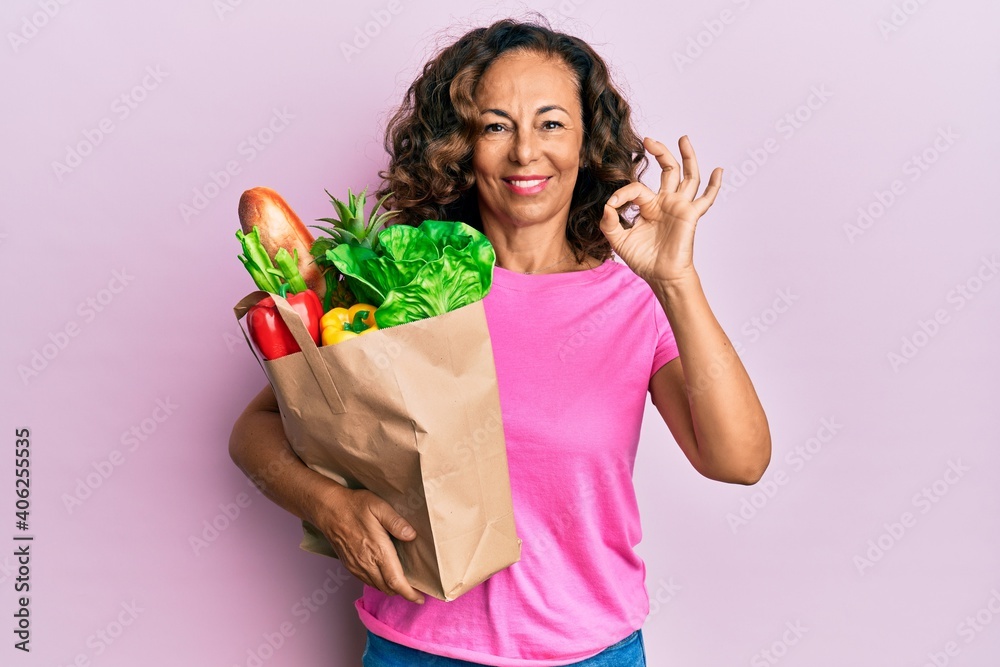 Middle age hispanic woman holding paper bag with bread and groceries doing ok sign with fingers, smiling friendly gesturing excellent symbol