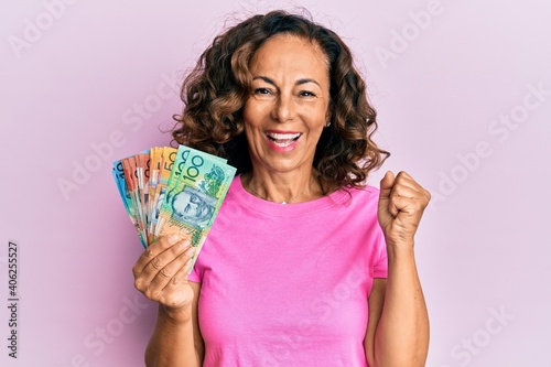 Middle age hispanic woman holding australian dollars screaming proud, celebrating victory and success very excited with raised arm