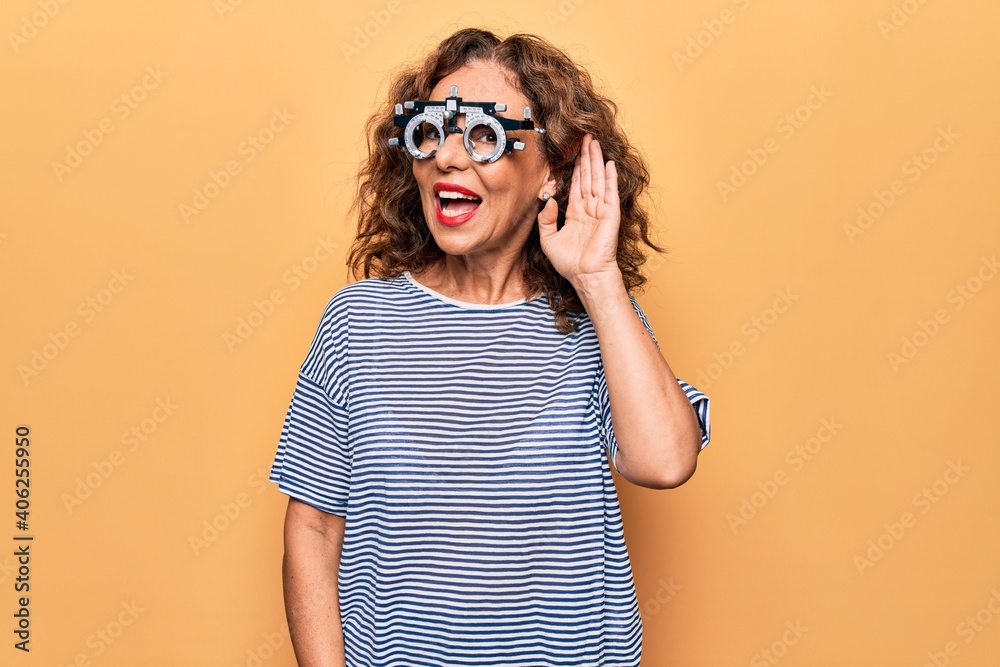 Middle age beautiful woman controlling vision using optometry glasses over yellow background smiling with hand over ear listening and hearing to rumor or gossip. Deafness concept.