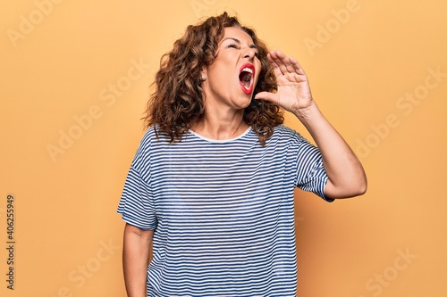 Middle age beautiful woman wearing striped t-shirt standing over isolated yellow background shouting and screaming loud to side with hand on mouth. Communication concept.