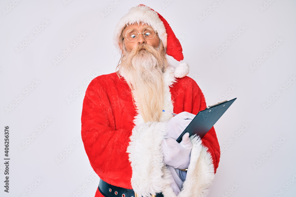 Old senior man with grey hair and long beard wearing santa claus costume writting list making fish face with mouth and squinting eyes, crazy and comical.