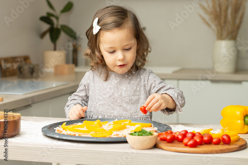 Cute little girl 2-4 in gray dress cooking pizza in kitchen. Kid arranges ingredients on pizza base