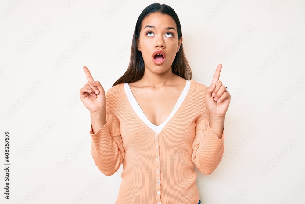 Young beautiful latin girl wearing casual clothes amazed and surprised looking up and pointing with fingers and raised arms. Young beautiful latin girl wearing casual clothes amazed and surprised looking up and pointing with fingers and raised arms.