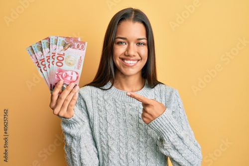 Beautiful hispanic woman holding 100 new zealand dollars banknote smiling happy pointing with hand and finger
