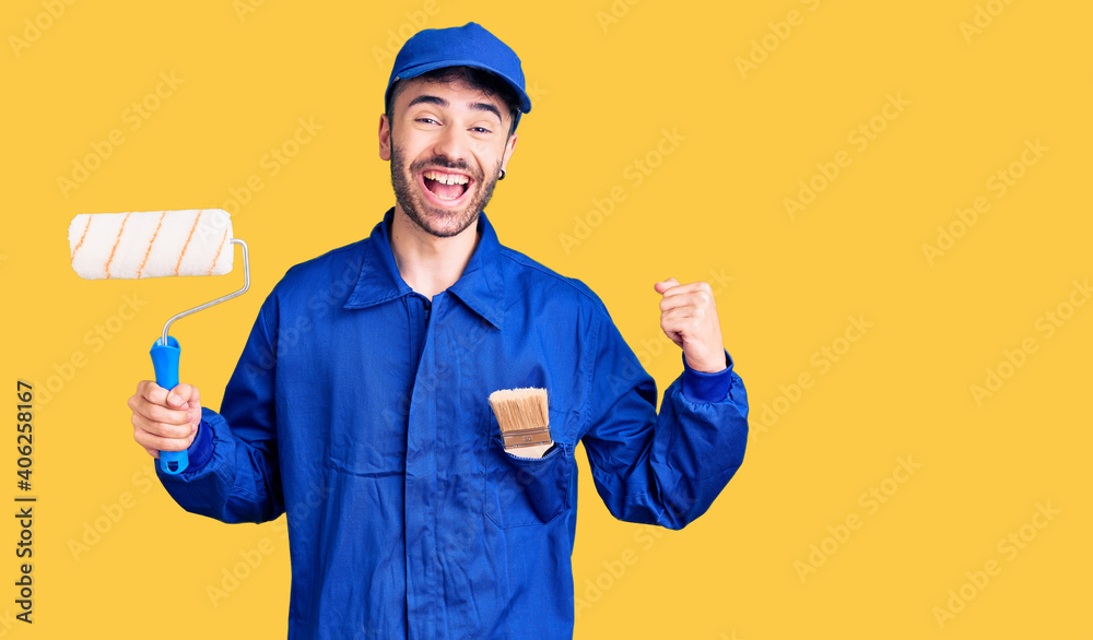 Young hispanic man wearing painter uniform holding roller screaming proud, celebrating victory and success very excited with raised arms