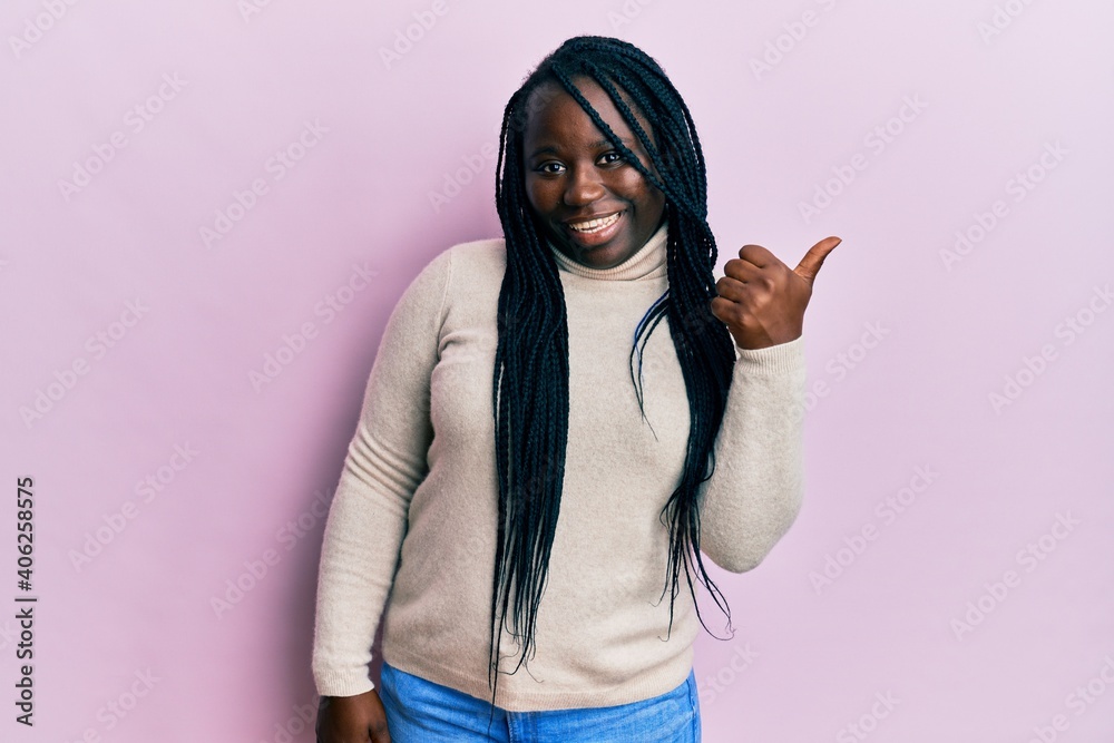 Young black woman with braids wearing casual winter sweater smiling with happy face looking and pointing to the side with thumb up. Young black woman with braids wearing casual winter sweater smiling with happy face looking and pointing to the side with thumb up.