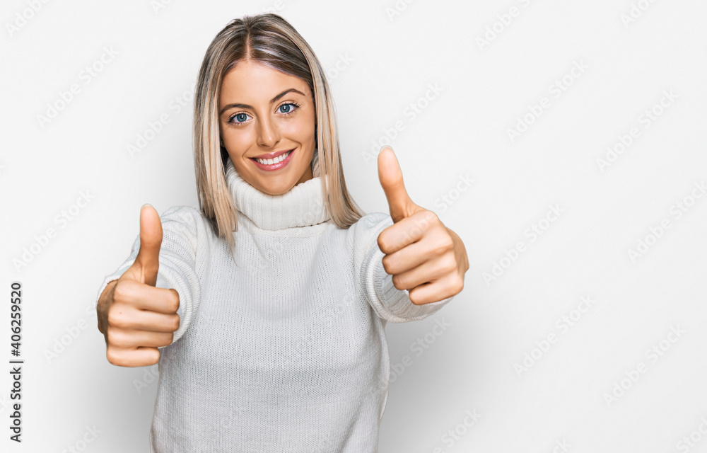 Beautiful blonde woman wearing casual turtleneck sweater approving doing positive gesture with hand, thumbs up smiling and happy for success. winner gesture.