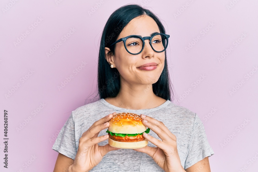 Beautiful young woman eating tasty hamburger smiling looking to the side and staring away thinking.