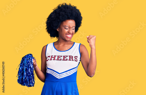 Fotografie Young african american woman wearing cheerleader uniform holding pompom very happy and excited doing winner gesture with arms raised, smiling and screaming for success