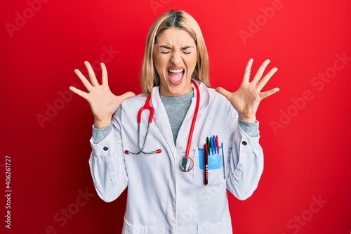 Young caucasian woman wearing doctor uniform and stethoscope celebrating mad and crazy for success with arms raised and closed eyes screaming excited. winner concept