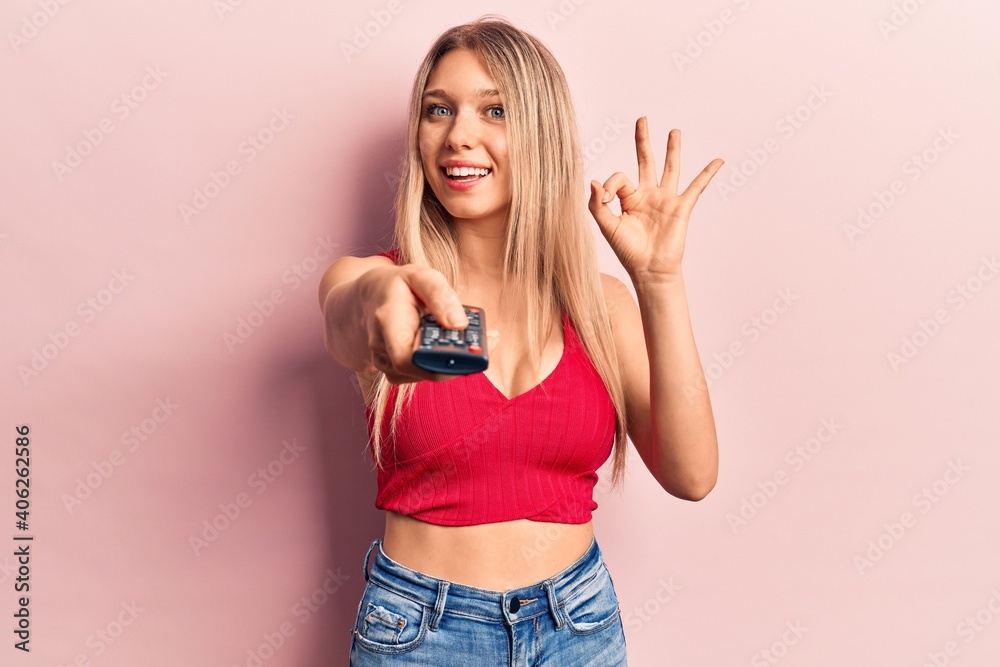 Young blonde woman holding television remote control doing ok sign with fingers, smiling friendly gesturing excellent symbol Young blonde woman holding television remote control doing ok sign with fingers, smiling friendly gesturing excellent symbol