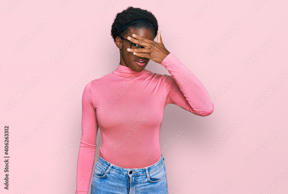 Young african american girl wearing casual clothes and glasses peeking in shock covering face and eyes with hand, looking through fingers with embarrassed expression. Young african american girl wearing casual clothes and glasses peeking in shock covering face and eyes with hand, looking through fingers with embarrassed expression.