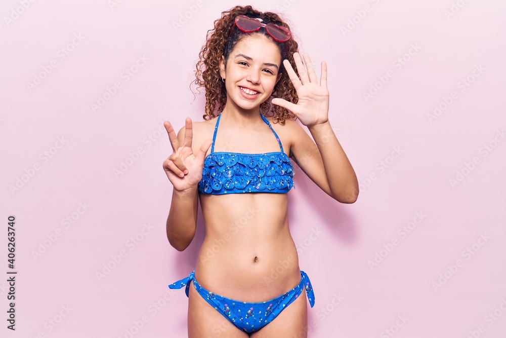 Naklejka premium Beautiful kid girl with curly hair wearing bikini and sunglasses showing and pointing up with fingers number eight while smiling confident and happy.